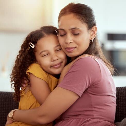 Mixed race single mother and daughter hugging in home living room. Smiling hispanic girl embracing and bonding with single parent in lounge. Happy affectionate woman and child together on weekend