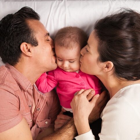 Couple of new parents lying in a bed with her baby girl and kissing her both at the same time